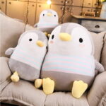 Three chubby penguin plushies are displayed on a couch. The plushies are gray with white stripes and yellow feet.