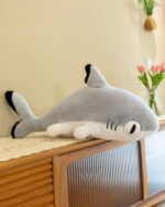 A gray giant shark plushie, Fins the Chonky Shark, lies on a wooden cabinet.