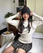 Woman wearing a beige korean aesthetic sweater with a bowknot print, sitting on a bed.