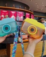 Two Kawaii Camera Portable Drinking Bottles, teal and yellow, held by hands with a colorful strap.