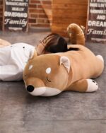 Kawaii lazy shiba plushie resting on a bed, with a person lying down with it.