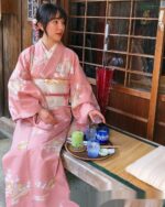 A woman wearing a traditional japanese kimono sits with tea cups on a tatami mat.