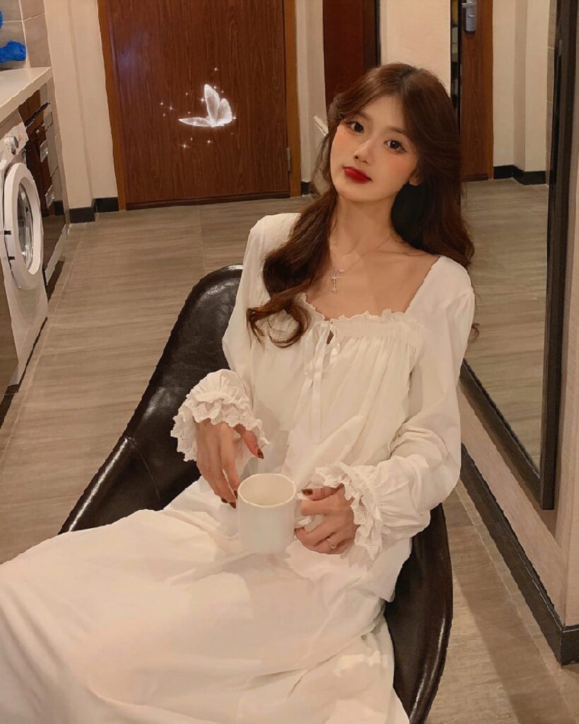Woman wearing a white soft girl aesthetic nightdress, holding a mug in a room.