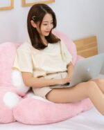Pink kawaii neko pillow displayed on a bed with a person working on a laptop.