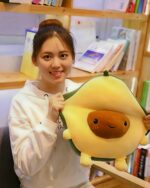 A brown kawaii avocado plushie is held by a woman in a bookstore.