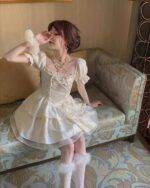 Cream vintage aesthetic dress with square collar, layered fabric, and puffed sleeves, seated on a patterned sofa.