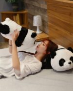 A large, fluffy kawaii panda plushie lies on a bed with a person.