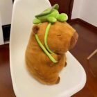 A brown capybara plushie wearing a green turtle accessory, sitting on a white chair.