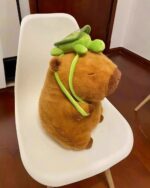 A brown capybara plushie wearing a green turtle accessory, sitting on a white chair.