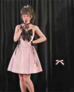 Young woman wearing a pink kawaii goth dress with a black bow and lace details, standing in front of black curtains.