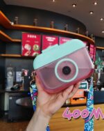 Hand holding a kawaii camera bottle, teal and pink design, portable water bottle.