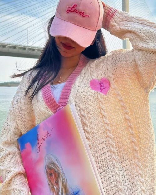 Embroidered cardigan sweater in a beige knit with heart embroidery, styled with a pink hat and book.