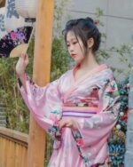 Woman wearing a pink short floral kimono with floral and bird patterns, holding a fan.