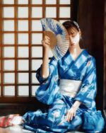 Woman wearing a deep blue yukata with floral patterns, holding a fan indoors.