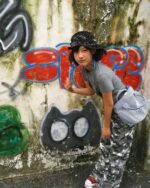 Model wearing a retro grunge t-shirt with camouflage pants and a hat, posing against a graffiti wall.