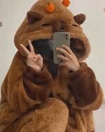 Person wearing a brown capybara hoodie, holding a phone and making a peace sign.