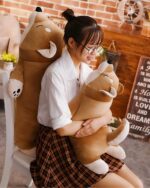A brown lazy shiba plushie is held by a person in a chair.