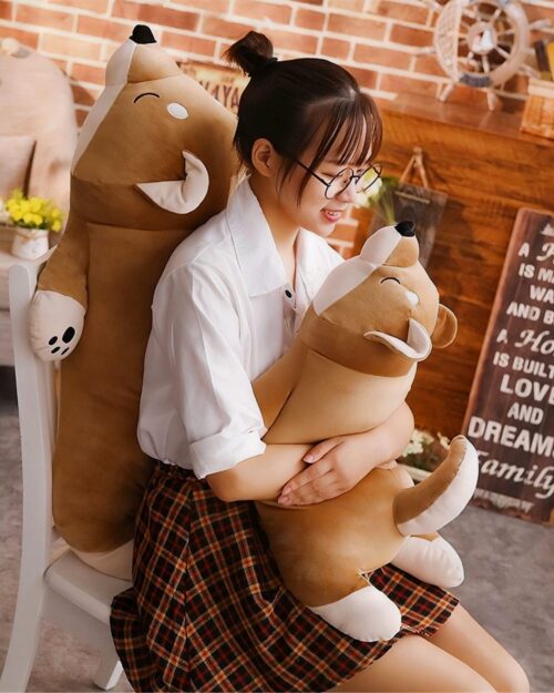 A brown lazy shiba plushie is held by a person in a chair.