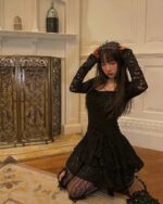 Woman wearing a black lace gothic aesthetic dress, long sleeves, and crown. Decorative fireplace in background.