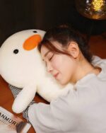 Close-up of a white kawaii duck plushie, Winston, resting on a bed with a person sleeping.