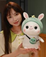 Cotton the Adventurous Rabbit, a kawaii bunny plushie, holds a green frog pendant and wears a cute hat.