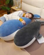 A large kawaii whale plushie, Bubbles, resting on a bed with a person. It's a soft, blue-grey plush toy.