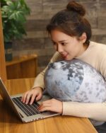 A woman using a laptop with a yuki the seal plush pillow resting on it.