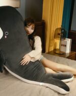 A gray kawaii whale plushie lies on a bed, hugged by a person.