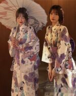 Two women wearing a light purple yukata with floral and geometric patterns, holding parasols.