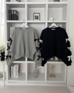 Two black and gray bowed pullover sweaters hang on white hangers, displayed on a shelf.