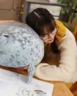 A grey Yuki the Seal Plush is resting on a woman's lap, alongside an open book.