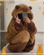 A brown Capyvara Hooded Coat, featuring a hood and paws, shown with a phone and hand gestures.