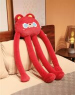 A red long-legged bear plushie resting on a bed with pillows.