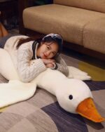 A large, white big goose plush toy lies on a rug with a person.