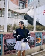 Woman wearing a dark blue bowed pullover sweater with white bows, styled with a white skirt and bag.