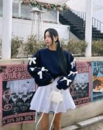 Woman wearing a navy bowed pullover sweater layered over a white skirt, outdoors.