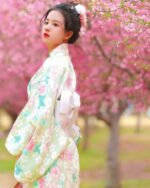 A woman wearing a light green japanese floral kimono outdoors among cherry blossoms.
