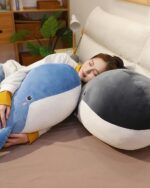 Blue and grey kawaii whale plushie, Bubbles, resting on a bed alongside a person.