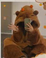 Person wearing a brown capybara hoodie and holding a smartphone.