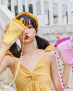 A pink and purple kawaii fruit bottle with a strap, being held by a woman outdoors.