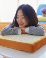 Cute toast cat bed plush pillow with a woman resting her hand on it. It is a soft, yellow-brown color.