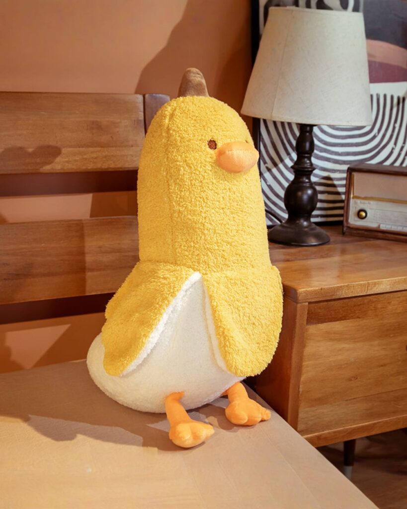 A cheerful banana duck plushie, featuring soft yellow and white fur, orange feet, and a brown beak.