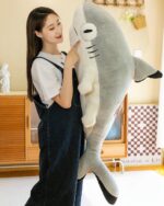 Large, plush giant shark plushie, Fins the Chonky Shark, resting in a woman’s arms.