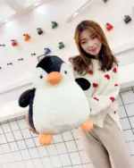 A large, black and white chonky penguin plushie sits next to a woman holding it.