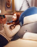 A blue kawaii whale plushie lies on a bed with a woman reading on her phone.