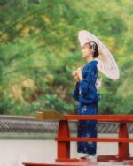 Woman wearing a deep blue yukata stands on a bridge, holding an umbrella in a Japanese garden.
