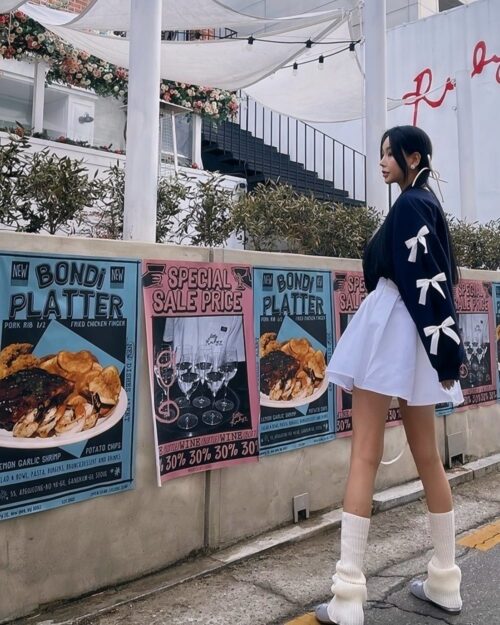 Woman wearing a blue bowed pullover sweater, outdoors with food stall signage.