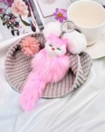 Pink fox plush keychain hangs on a gray knitted ring, with a keyring and cup in the background.