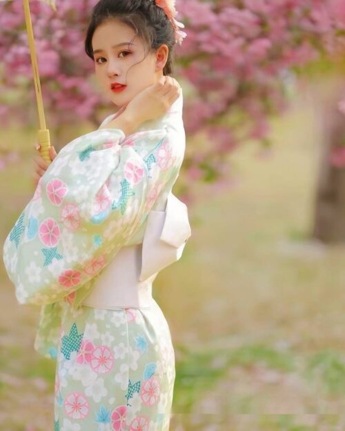 Woman wearing a light blue japanese floral kimono outdoors among cherry blossoms.