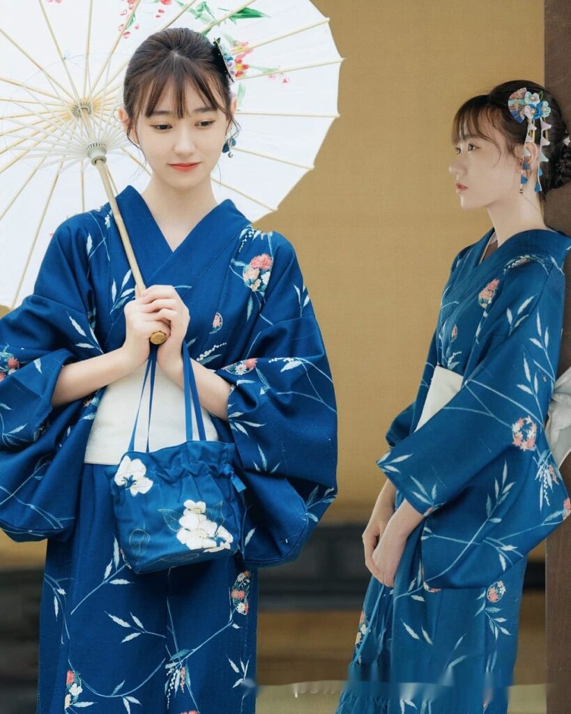 Two women wearing a deep blue yukata with floral pattern, holding umbrellas.